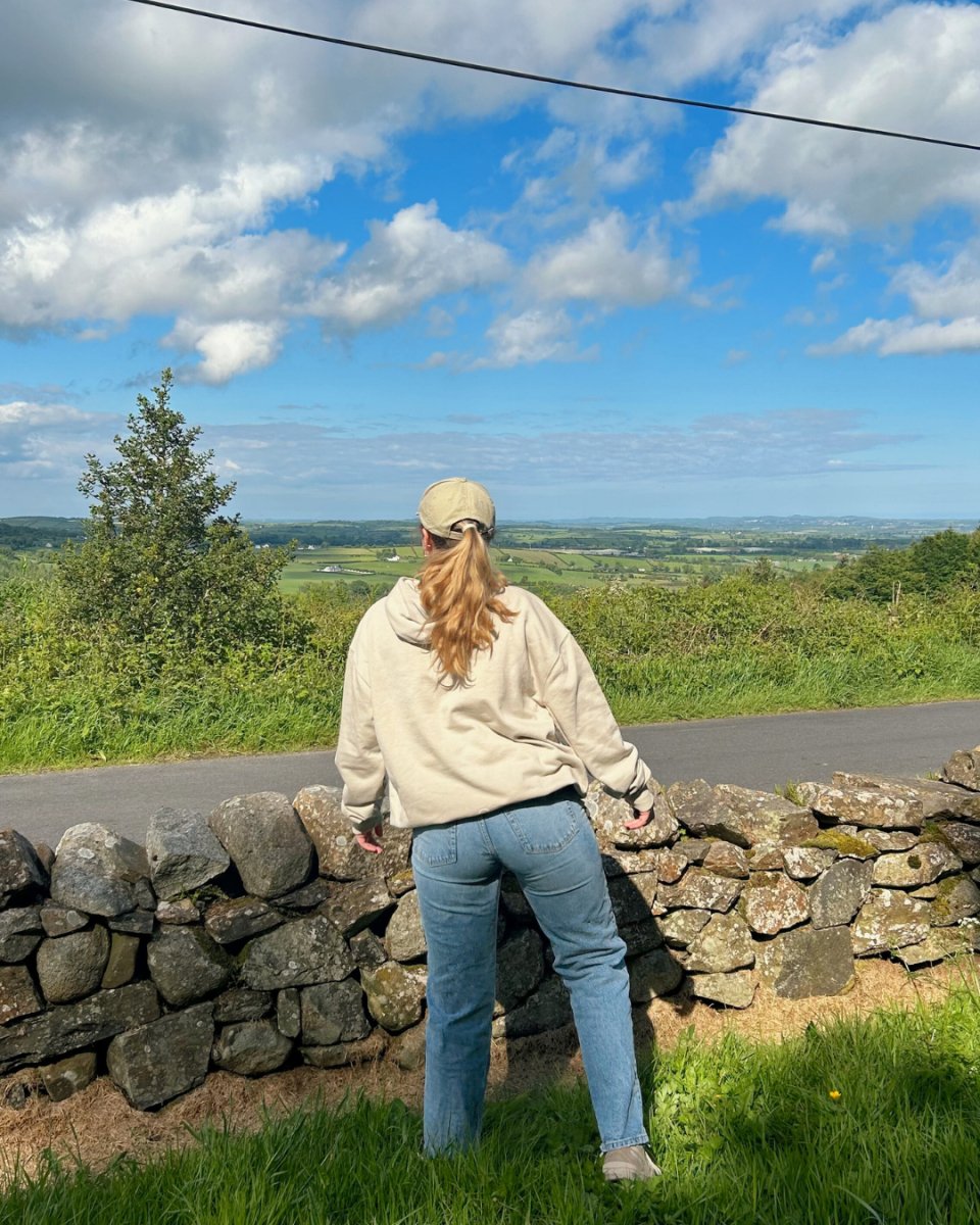 A person wearing a beige hoodie and light blue jeans, with a beige cap, standing beside a stone wall and looking at a scenic view. Vegan clothing from Compassion Threads.