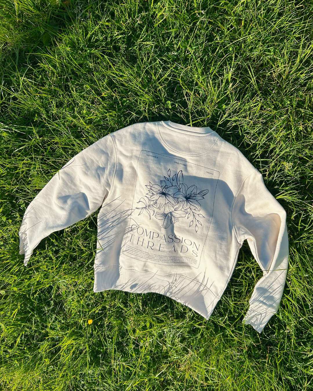 A beige sweatshirt with a floral design and the text 'Compassion Threads' displayed on the back, laid out on lush green grass in a natural setting. conscious clothing and ethical fashion options available from Compassion Threads