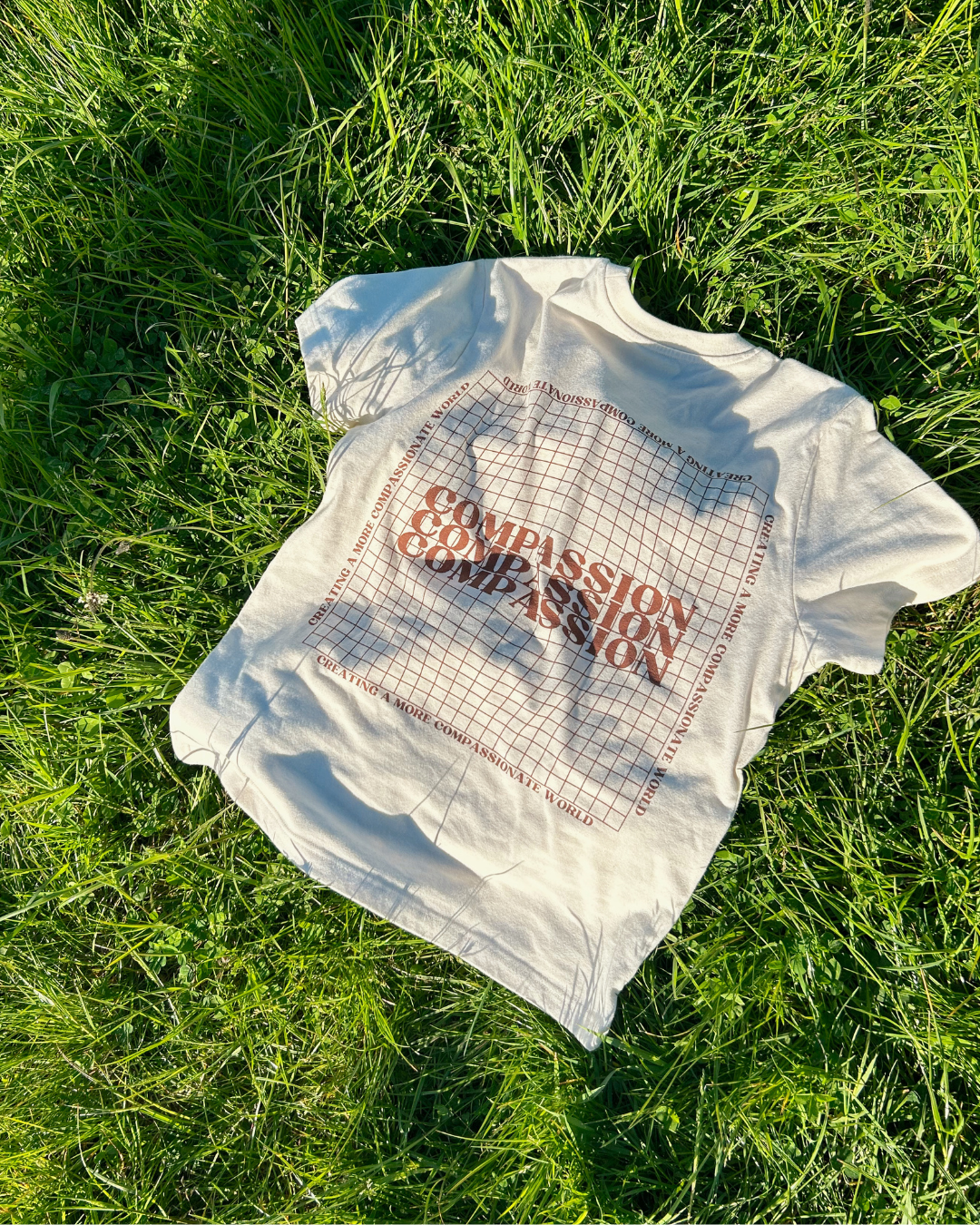 A beige t-shirt with the word 'Compassion' repeated in a grid pattern and the phrase 'Creating a More Compassionate World' around the border, laid out on lush green grass. fashion with a cause, ethical clothing from Compassion Threads.