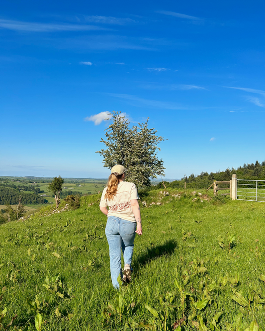 A person wearing a beige t-shirt with a grid pattern and the word 'Compassion' on the back, paired with light blue jeans and a beige cap, walking through a grassy field under a clear blue sky. sustainable premium loungewear from Compassion Threads.