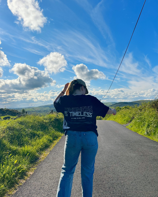A person wearing a navy t-shirt with the text 'Kindness is Timeless' and 'Be Kind to All Kind' on the back, paired with light blue jeans and a beige cap, walking along a paved road with a scenic view. Sustainable fashion from Compassion Threads.