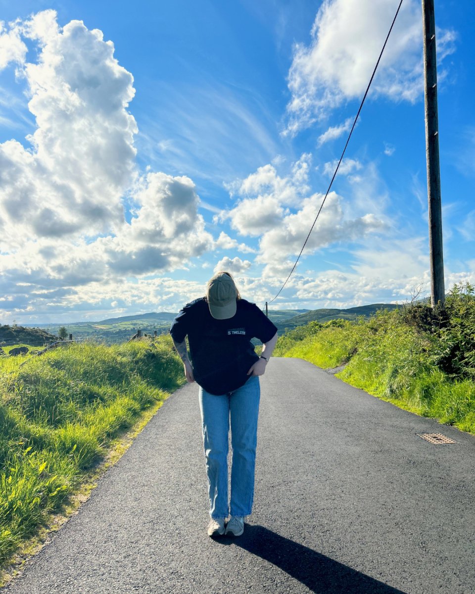 A person wearing a navy t-shirt with the text 'Kindness is Timeless' and 'Be Kind to All Kind' on the back, paired with light blue jeans and a beige cap, standing on a paved road with a scenic view. Sustainable fashion from Compassion Threads.