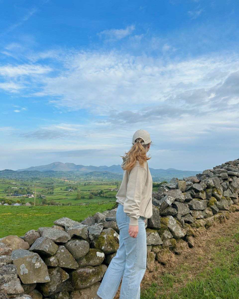 A person wearing a beige hoodie and light blue jeans, with a beige cap, standing beside a stone wall and looking at the scenic landscape. Sustainable premium loungewear from Compassion Threads.