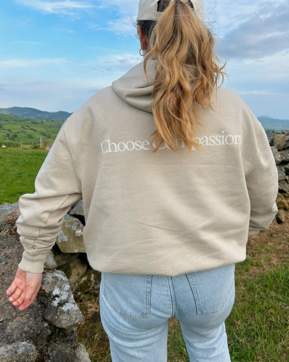 A person wearing a beige hoodie with the text 'Choose Compassion' on the back, paired with light blue jeans and a beige cap, standing beside a stone wall with a scenic view sustainable fashion, ethical, eco-friendly vegan clothing