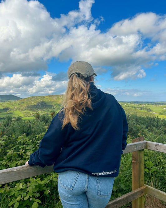 A person wearing a black hoodie with the text 'Compassionate World' on the navy, paired with light blue jeans and a beige cap, standing by a wooden fence and looking at a scenic view. compassionate fashion and ethical hoodies from Compassion Threads