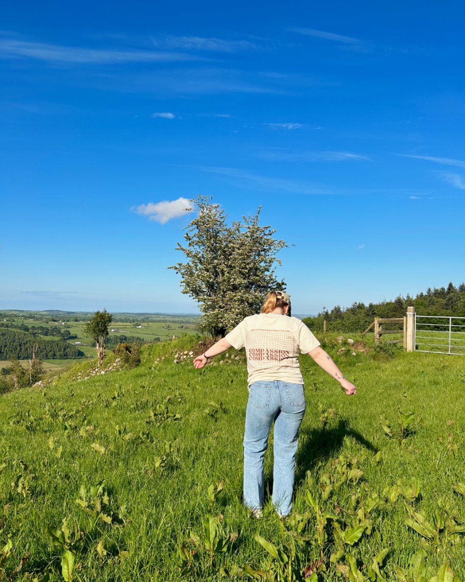 A person wearing a cream t-shirt with a grid pattern and the word 'Compassion' on the back, paired with light blue jeans, standing in a grassy field with a clear blue sky and a view of green hills.