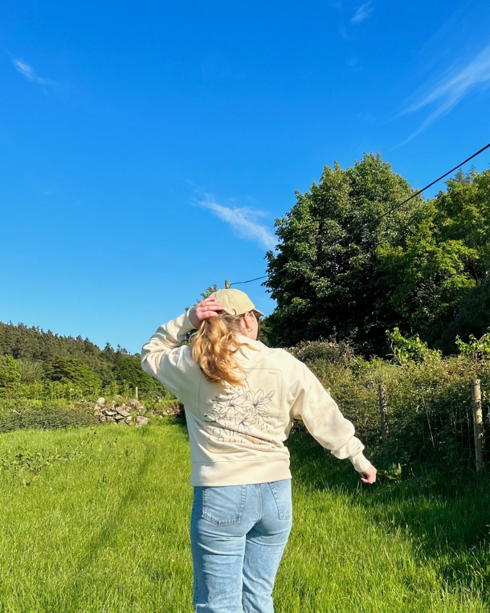 A person wearing a beige hoodie with a floral design and the text 'Kindness Over Everything' on the back, paired with light blue jeans and a beige cap, walking through a grassy field under a clear blue sky. vegan and ethical clothing