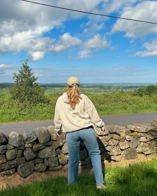 A person wearing a beige hoodie and light blue jeans, with a beige cap, standing beside a stone wall and looking at a scenic view. Vegan clothing from Compassion Threads.