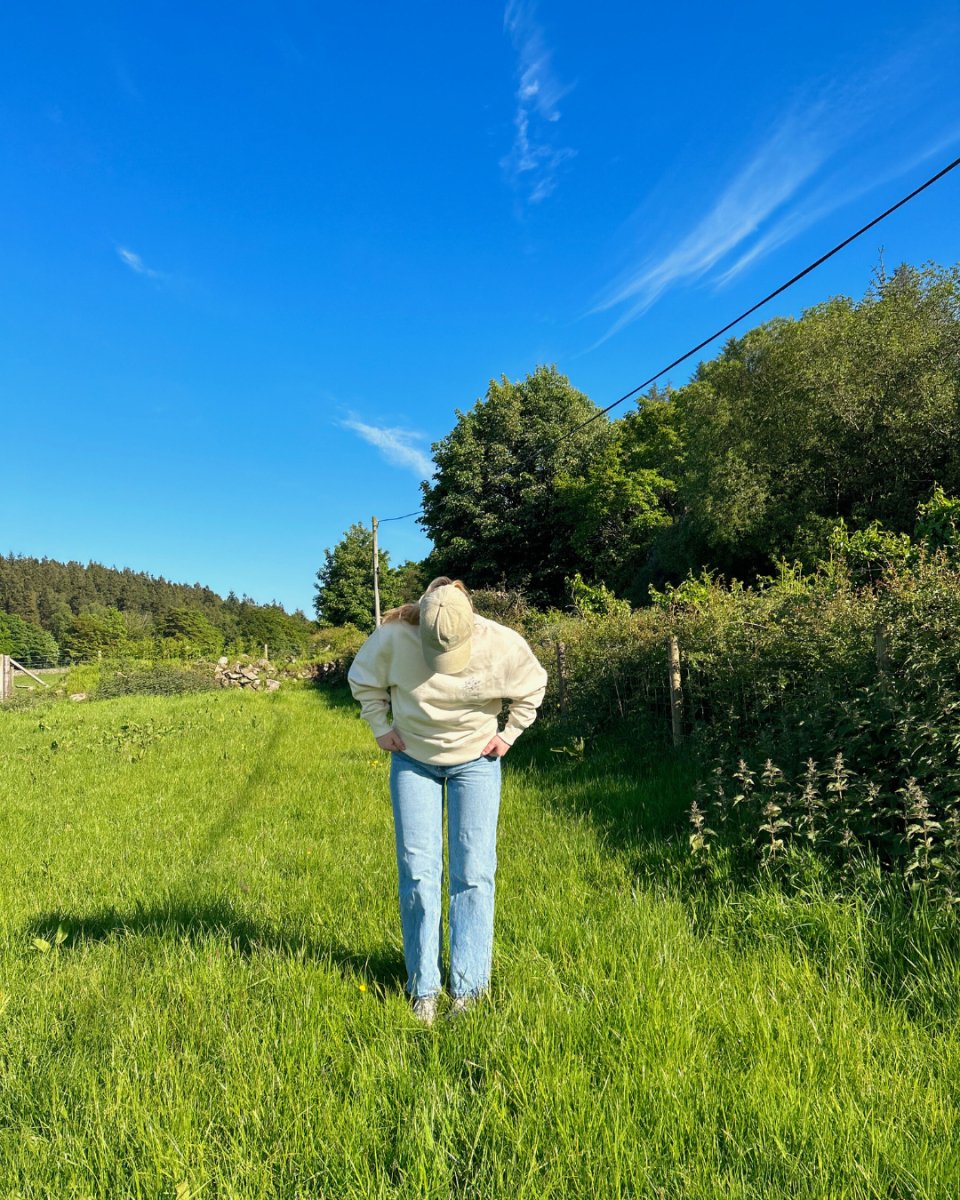 A person wearing a beige sweatshirt and blue jeans, with a beige cap, standing in a grassy field and looking down. vegan and ethical clothing 