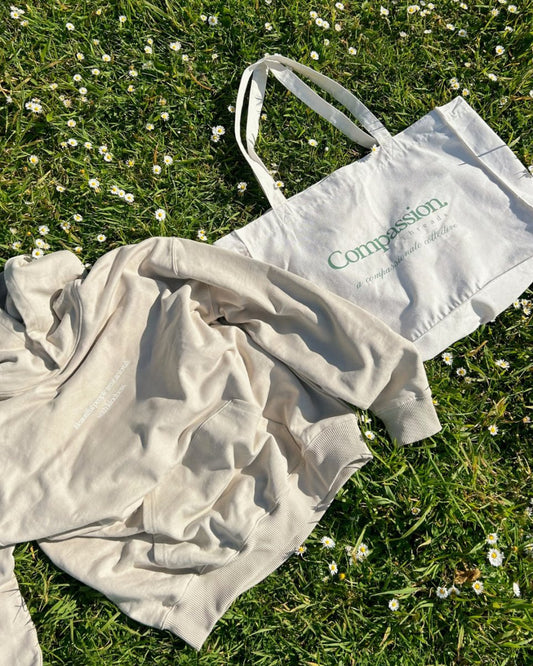 A beige hoodie and a white tote bag with the text 'Compassion' and 'a compassionate collective' displayed on the grass with small white flowers. Ethical hoodies and sustainable, cruelty-free fashion.